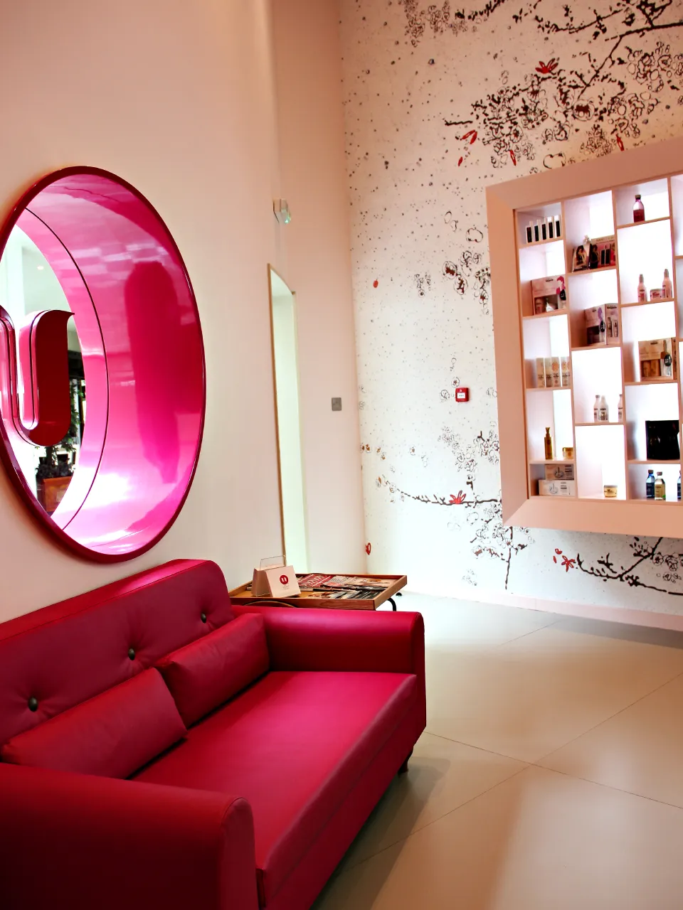 Beauty salon waiting lounge in Dubai with red sofa, circular red mirror, branch wall graphics, magazine tray, and illuminated product display shelves
