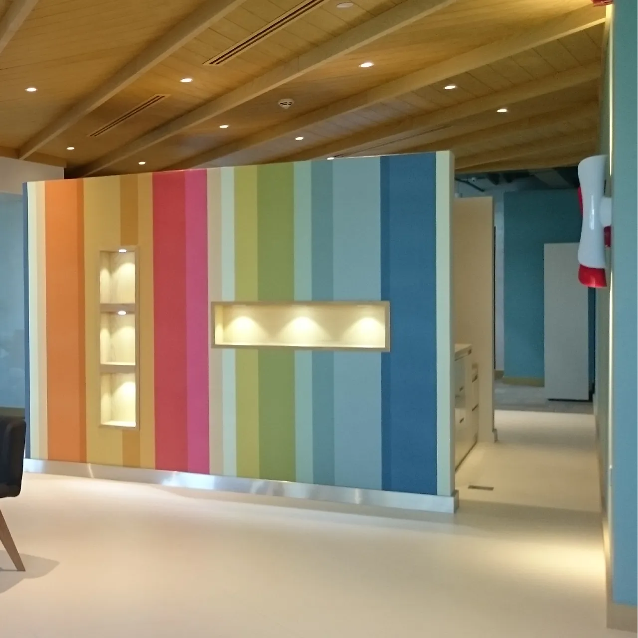 Small factory office design with a multicolored vertical feature wall, illuminated display niches, pale flooring, and timber ceiling