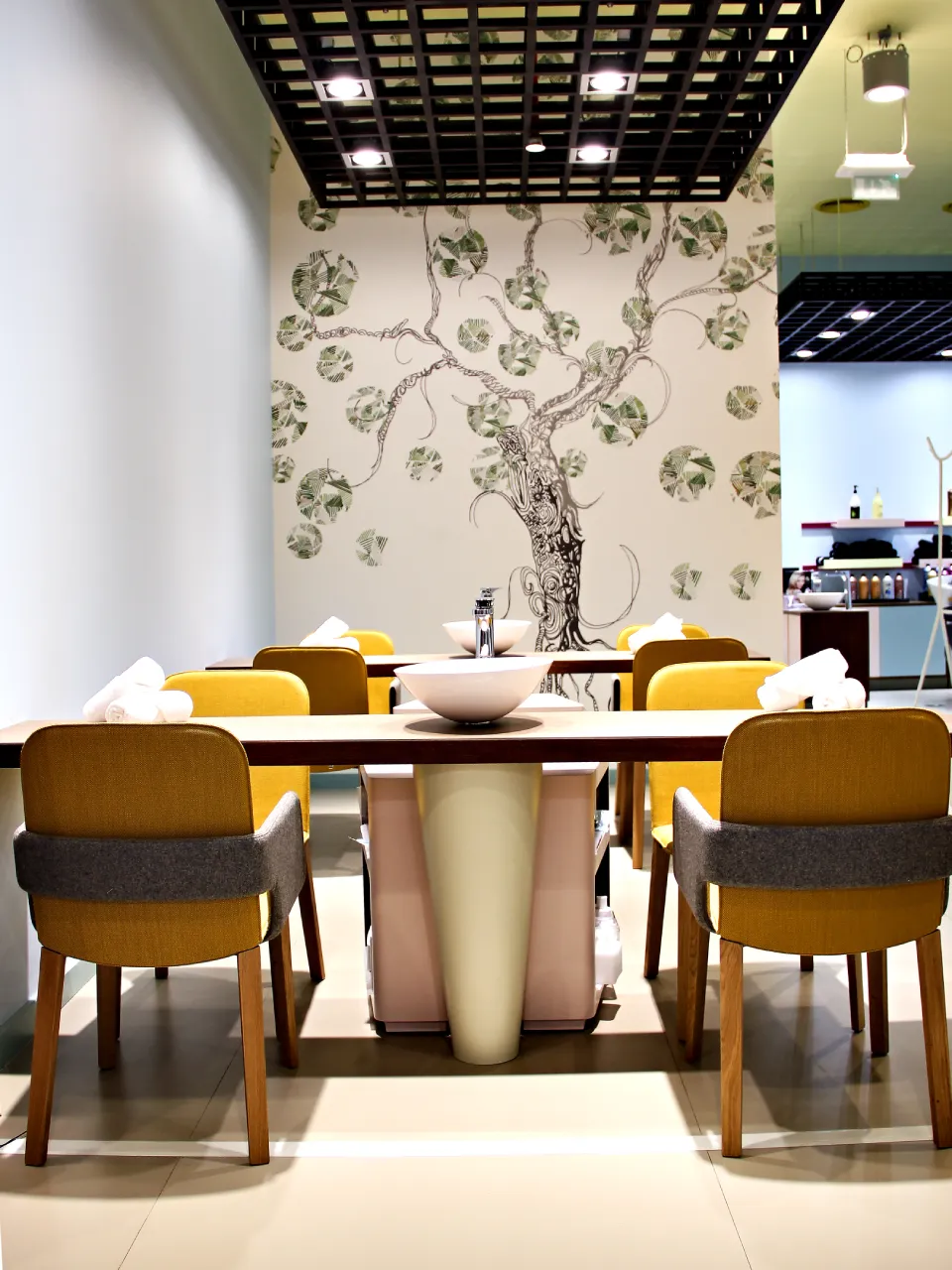 Dubai beauty salon manicure area with yellow chairs, black grid ceiling, white service tables, towels, and illustrated tree mural