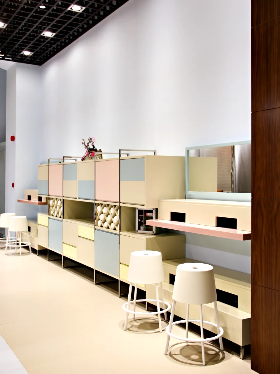 Dubai beauty salon retail area with pastel modular cabinets, open product shelves, long counter, white stools, black grid ceiling, and warm wood panel