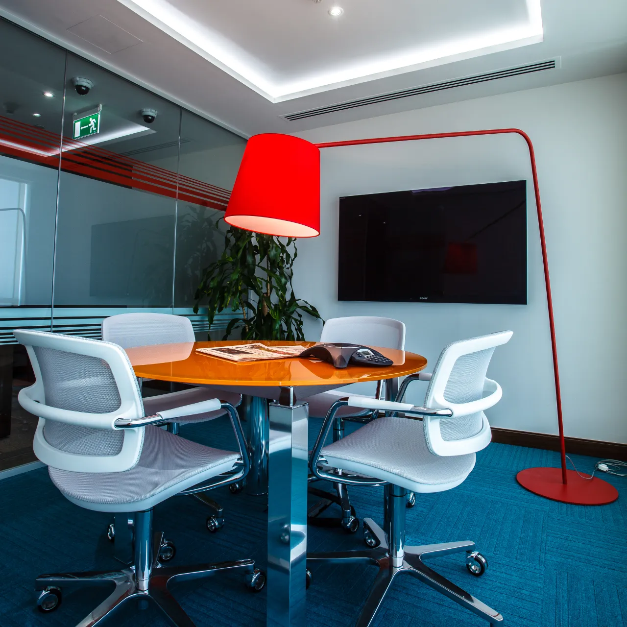 IT company office interior with a circular collaboration table, sculptural red lamp, glass partition, television, and concealed ceiling lighting